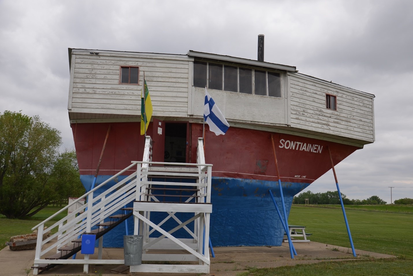 Pictured is a photo of the Sukanen Ship Pioneer Museum. The building is actually a ship on a concrete base supported by metal poles, refashioned into a building for the museum. The ship is white, red, and blue in thick stripes going top to bottom, and the entrance is on the second floor; there is a white staircase leading to the door. The front door is flanked by the Saskatchewan flag on the left and the Finnish flag on the right. On the side of the ship is the name 'Sontiainen' in white text. In the background is green grass and a cloudy sky.