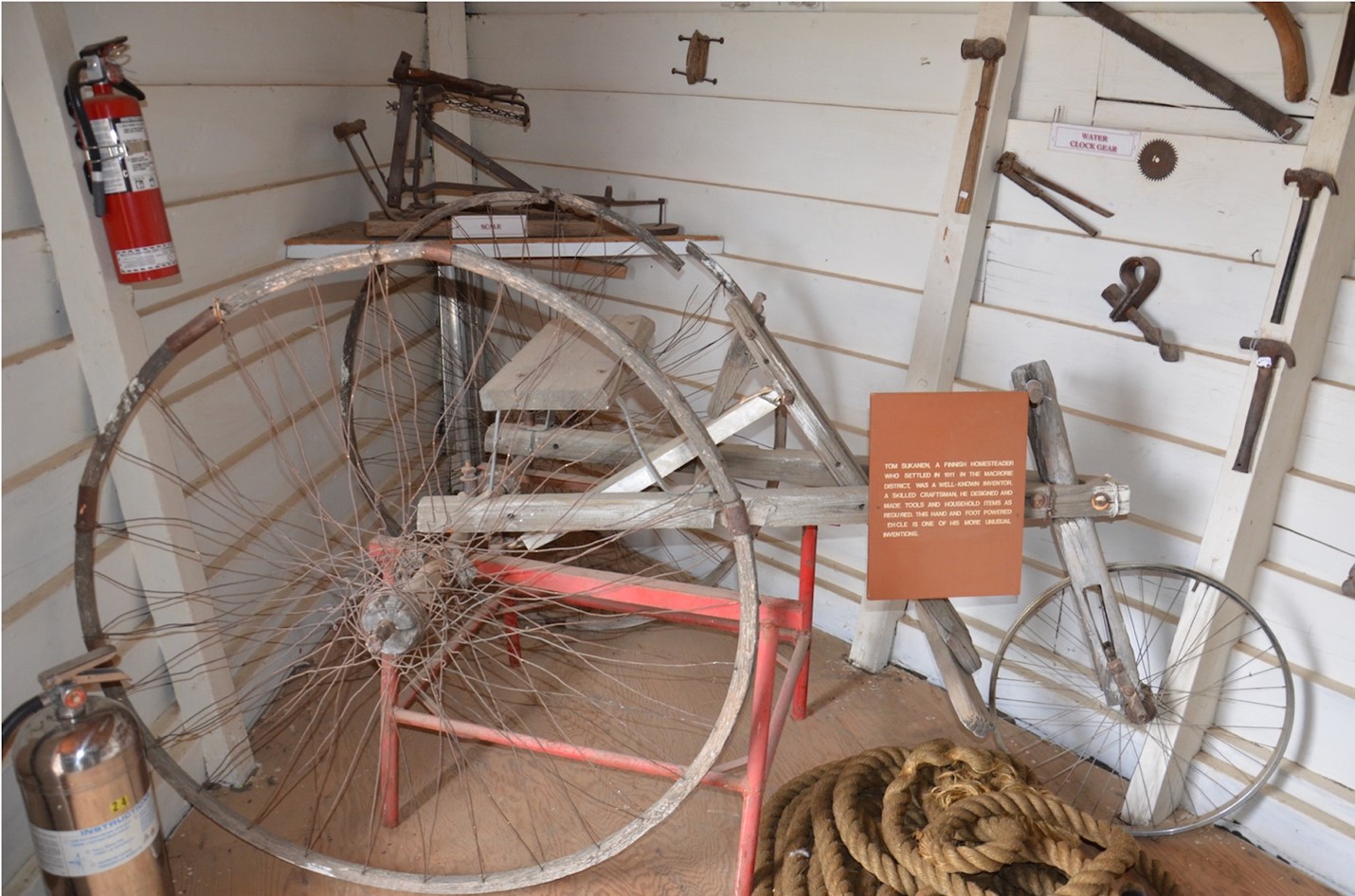 Pictured is a photo of a large tricycle, made by Tom Sukanen, being displayed in a corner of the Sukanen Ship Pioneer Village and Museum. The wall behind the tricycle is white shiplap and there are other assorted tools hanging from the wall. There is an orange sign attached to the tricycle which is illegible from the distance the photo was taken, presumably giving details about the item.