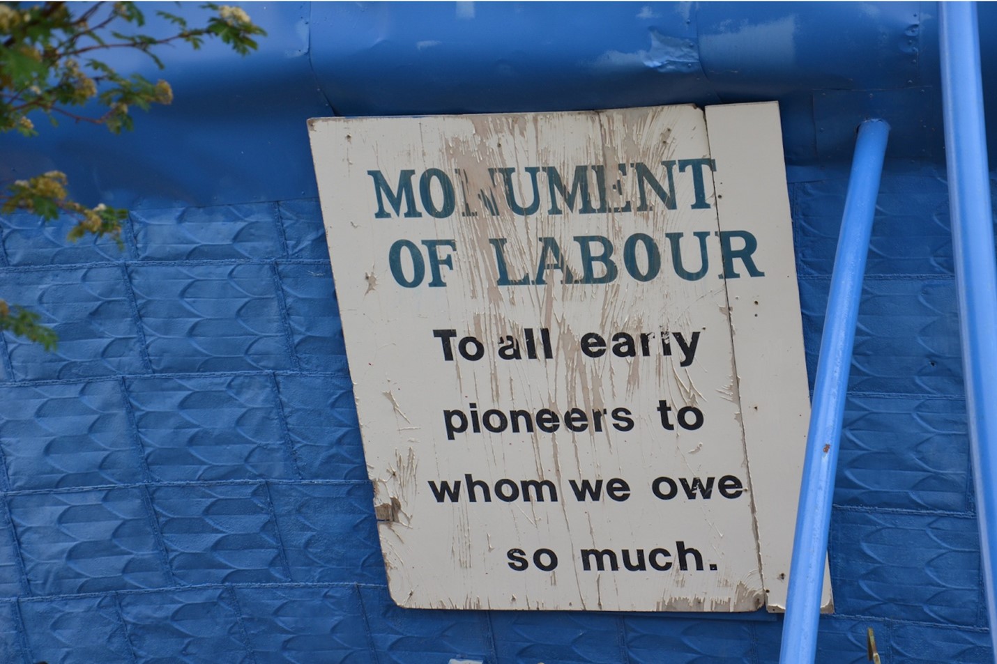Pictured is a photo of a sign attached to the side of the Sukanen Ship Pioneer Museum. The background is the royal blue of the ship, and the photo includes one of the matching blue metal support poles. The sign is white although the wood it is made of is weathered and partially showing through, and it reads 'MONUMENT OF LABOUR. To all early pioneers to whom we owe so much.' The text in all caps is dark blue and the rest is black. In the upper left corner of the image, a tree branch is intruding into the frame.