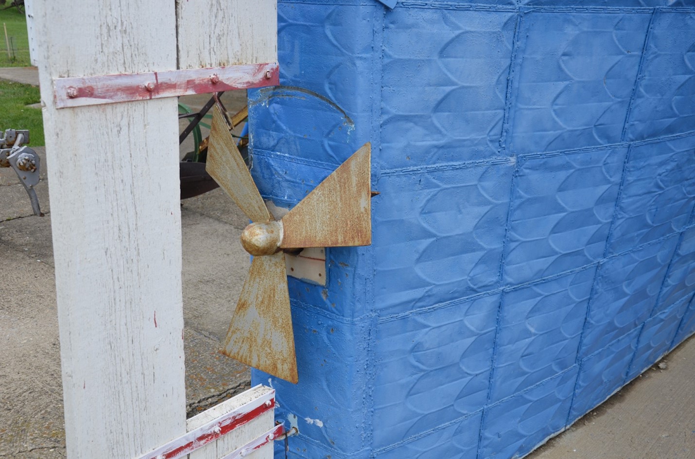 Pictured is a photo of part of the lower royal blue part of the Sukanen ship. The ship is made of some kind of metal which is arranged in squares and probably welded together. Within each square is a pattern of almost feather-shaped embossing or something similar. The ship sits on its concrete base and attached to the ship is a small, rusted propeller, previously silver or maybe white in colour. Protruding behind the propeller is a white wooden rectangle, with a cutout to make room for the propellar; it may be a rudder, or possibly protection for the propellar. The boards are attached to each other with red pieces of metal with rivets or screws and the whole thing is attached to the blue part of the ship at least at the bottom and possible further up, out of frame. There is a semi-circular scratch on the blue part of the ship that has rusted, where the white wooden piece looks like it has made contact. In the background, a small amount of green grass can be seen.