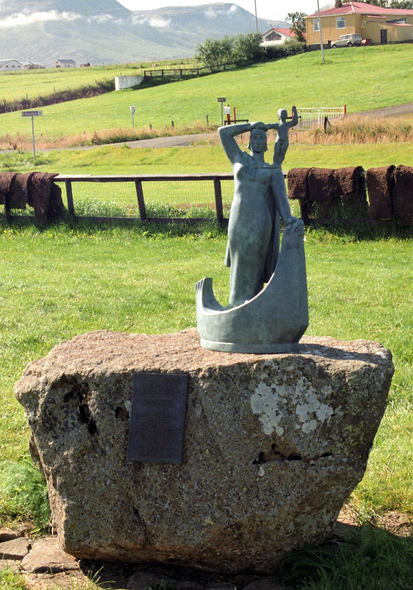 Pictured is The cast of Ásmundur Sveinsson’s Fyrsta hvíta móðirin í Ameríku. It is a small oxidized metal sculpture perched on top of a rock. The rock appears to be in a farmer's field, with a small woden fence behind it. Behind the fence is another field with a house in the background.