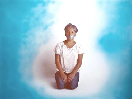 Anishinaabe artist Rebecca Belmore kneeling in an empty room, with duct tape covering her mouth.